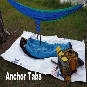 May include: A blue hammock with green accents is strung between two trees. A sleeping bag, a backpack, and a yellow foam pad are laid out on a white ground sheet. The text "Anchor Tabs" is visible in the lower left corner of the image.