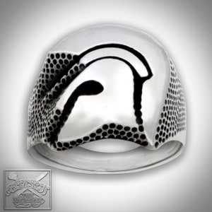 May include: A silver ring with a textured design featuring a Spartan helmet. The helmet is outlined in black and is centred on the ring.
