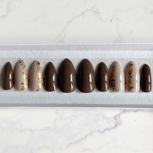 May include: Set of ten almond-shaped press-on nails with a brown and beige ombre design. Some nails have gold glitter accents.