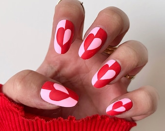 May include: Close-up of a hand with pink and red heart-shaped nail art. The nails are oval-shaped and painted with a pink and red color scheme, creating a heart design on each nail.