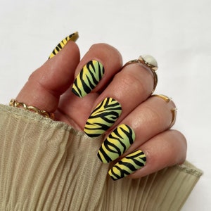 May include: Close-up of a hand wearing five long, yellow and black zebra print press-on nails. The nails are a matte finish and have a slightly rounded shape.