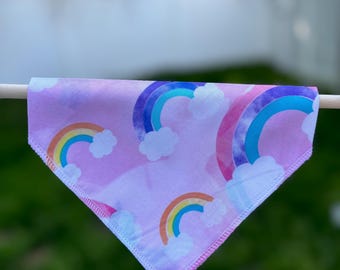 May include: A triangular, light purple bandana featuring a repeating pattern of colorful rainbows and fluffy white clouds. The rainbows have bands of red, orange, yellow, green, blue, and purple. The bandana is hanging on a wooden dowel.