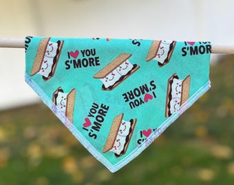 May include: A turquoise pet bandana with a repeating pattern of s'mores and the phrase "I ♥ You S'more." Each s'more has a smiling face. The bandana is trimmed with a white border and is hanging on a wooden dowel.