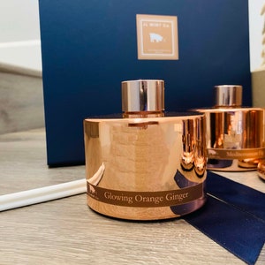 May include: Two copper-colored diffuser bottles with black labels. The bottle on the left reads "Glowing Orange Ginger". The bottle on the right reads "Mediterranean Fig".
