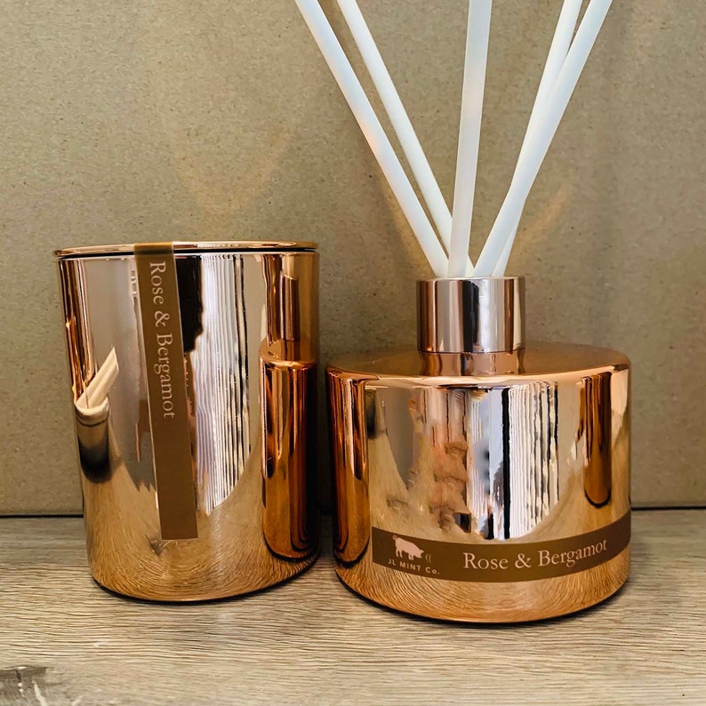 Puede incluir: Dos recipientes de vidrio de color oro rosa con el texto "Rose & Bergamot" en ellos. Un recipiente es una vela y el otro es un difusor con ca&ntilde;as blancas.