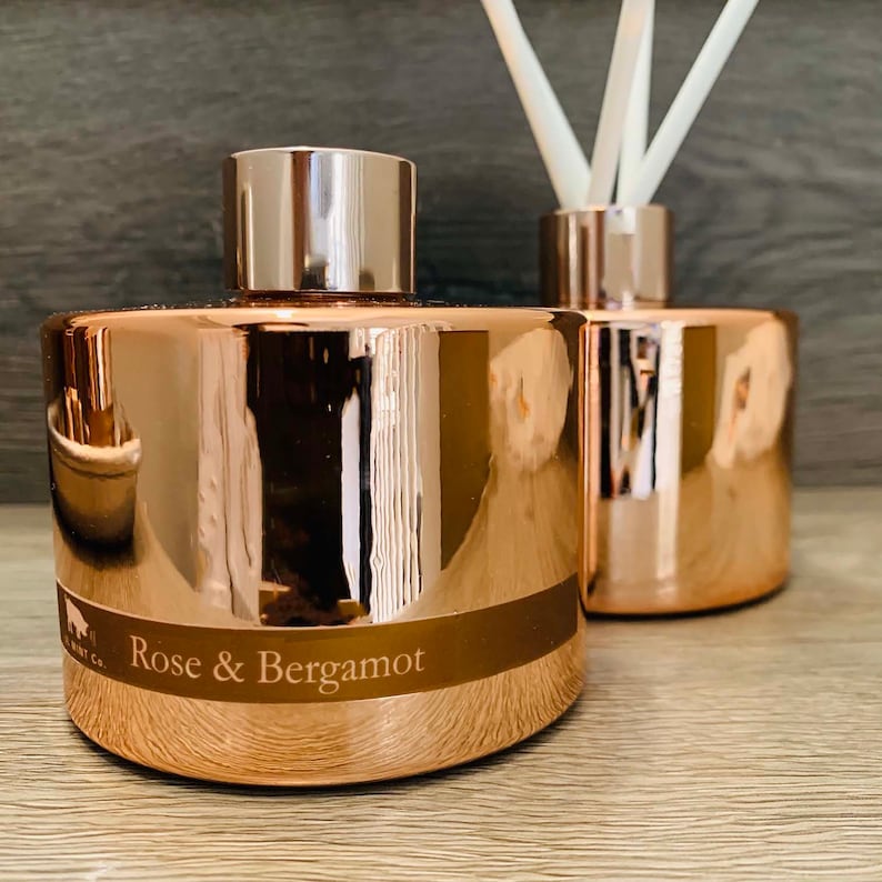 Puede incluir: Dos botellas difusoras de color oro rosa con una tapa de oro rosa y una etiqueta que dice "Rosa y Bergamota".