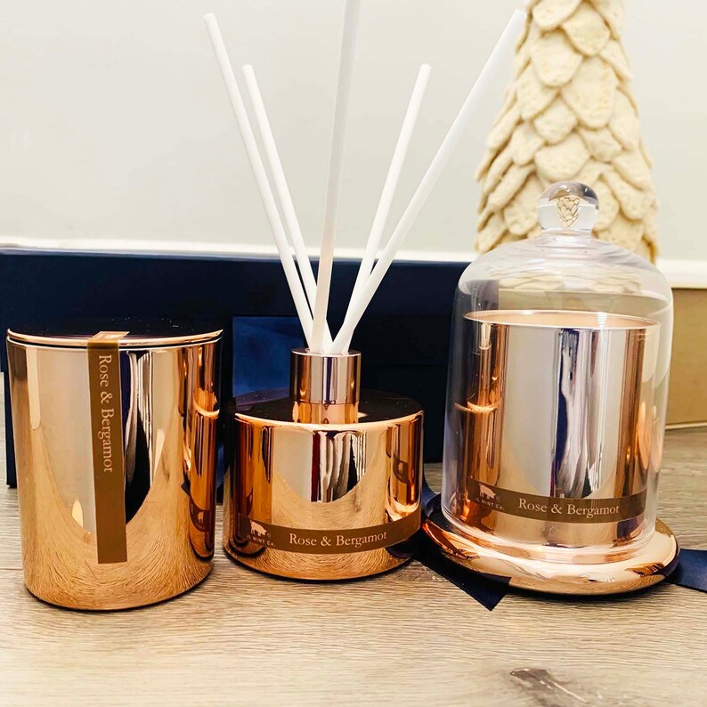 Puede incluir: Tres velas arom&aacute;ticas de color oro rosa con la etiqueta "Rose & Bergamota". Una vela est&aacute; en una campana de vidrio, otra est&aacute; en un frasco y otra est&aacute; en una botella de difusor con palitos.