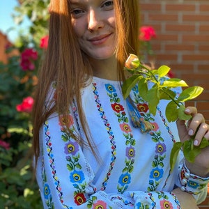 May include: A white embroidered blouse with a floral pattern in shades of blue, red, pink, and yellow. The blouse has long sleeves and a round neckline.