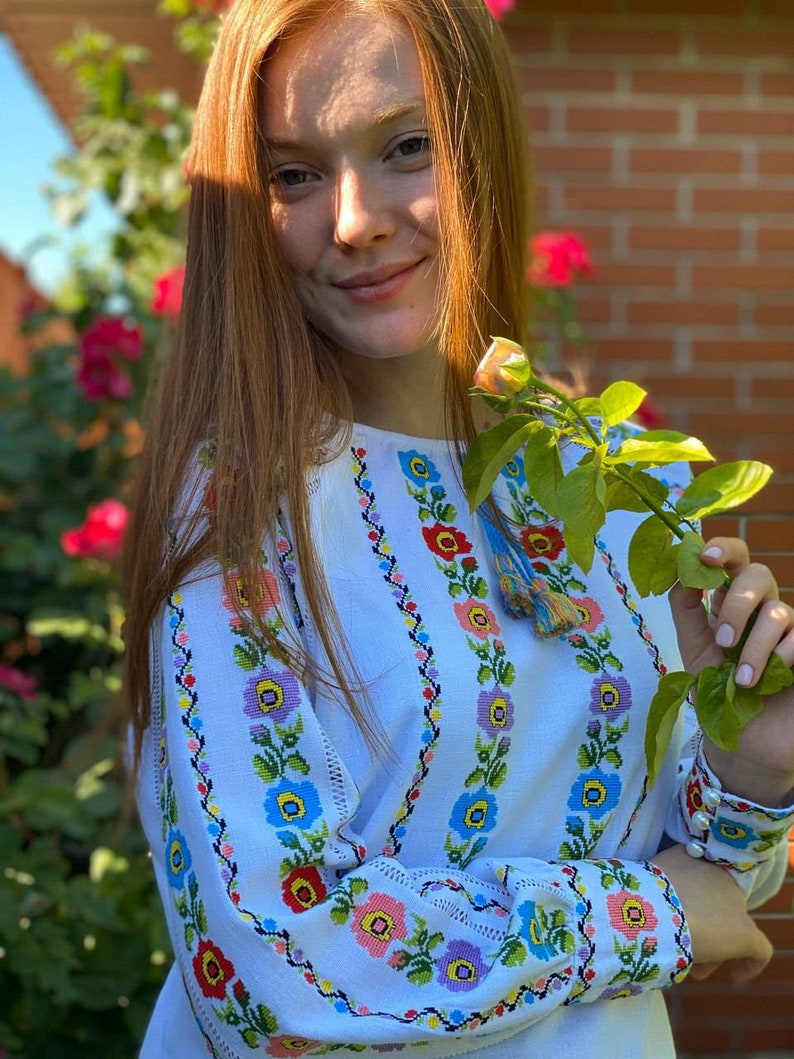 May include: A white embroidered blouse with a floral pattern in shades of blue, red, pink, and yellow. The blouse has long sleeves and a round neckline.