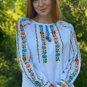 May include: White embroidered blouse with a floral pattern in shades of blue, pink, yellow, and green. The embroidery is on the sleeves and the front of the blouse.