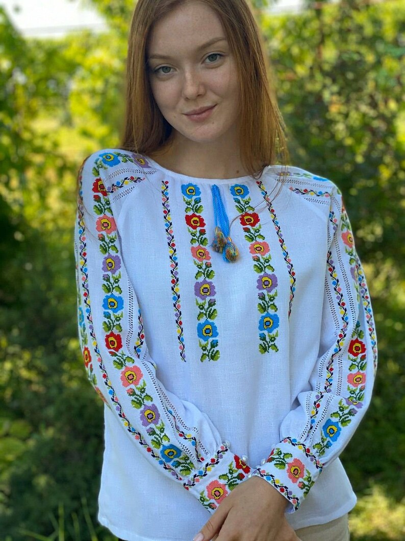 May include: White embroidered blouse with a floral pattern in shades of blue, pink, yellow, and green. The embroidery is on the sleeves and the front of the blouse.