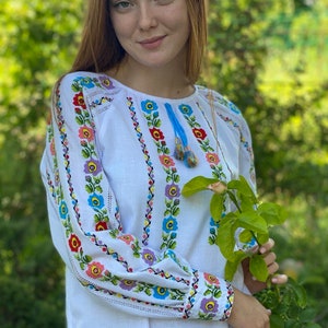 May include: A white linen blouse with colorful embroidered floral patterns. The embroidery features blue, pink, yellow, and green flowers. The blouse has long sleeves with a gathered cuff.
