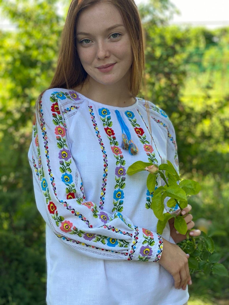 May include: A white linen blouse with colorful embroidered floral patterns. The embroidery features blue, pink, yellow, and green flowers. The blouse has long sleeves with a gathered cuff.