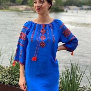 Puede incluir: Un vestido de lino azul con bordados rojos y blancos. El vestido tiene un corte holgado y un escote redondo. El bordado es de patrón geométrico.
