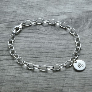 Puede incluir: Un brazalete de cadena de plata con un dije redondo que tiene la letra "M" grabada.