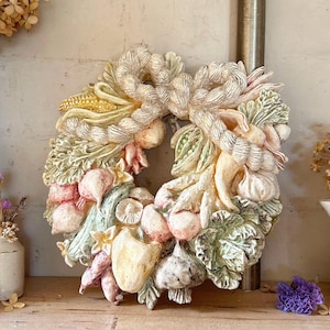 May include: A white ceramic wreath featuring a variety of sculpted vegetables and fruits, including corn, garlic, onions, and mushrooms. The wreath is adorned with a white ribbon and has a rustic, farmhouse style.