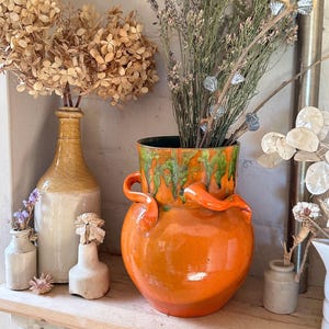 May include: Orange and green glazed ceramic vase with two handles. The vase is filled with dried flowers and is sitting on a wooden shelf.