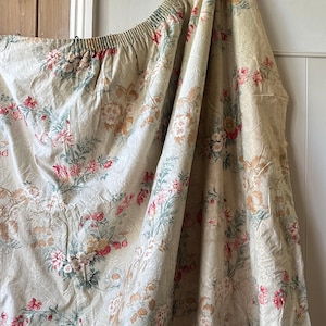 May include: A cream-coloured fabric featuring a floral pattern in pink, red, and green. The fabric is gathered at the top, possibly a curtain or drape. The floral design is detailed and has a vintage appearance.