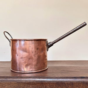 May include: A small copper pot with a long handle. The pot has a hammered finish and a rounded shape. The handle is made of iron and is attached to the pot with a rivet.
