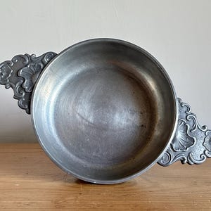 May include: A vintage pewter bowl with ornate, decorative handles. The bowl is round with a wide rim and a slightly textured surface. The handles feature a floral design. The bowl is displayed on a wooden surface.