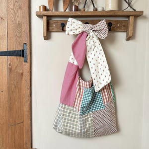 May include: A patchwork tote bag with a large bow, suspended from a wooden rack. The bag displays diverse fabric patterns, including red gingham, floral, and plaid designs. The bow is made of matching fabric.