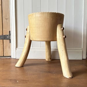 May include: A light-coloured wooden butcher block style stool with three legs. The stool has a round top and is supported by three angled legs. The legs are attached with small wooden pegs. The stool is made of unfinished wood.