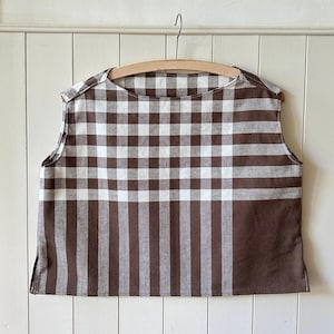 May include: A sleeveless top with a brown and white checkered pattern. The top is hanging on a wooden hanger against a white background. The fabric has a mix of vertical and horizontal lines.
