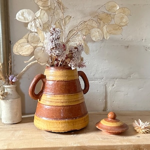 May include: A ceramic vase with a lid, featuring a brown and yellow striped design. The vase has two handles and is filled with dried flowers. A small, white vase with dried flowers is to the left.