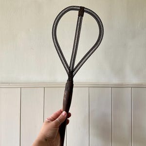 May include: An antique fly swatter with a dark wooden handle. The swatter head is made of a woven wire mesh in an oval shape. The handle is held by a person.