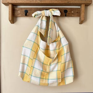 May include: A large, yellow and white plaid tote bag with a front pocket hangs from a wooden rack. The bag has a knotted handle and a teal grid pattern. The bag is made of a soft, lightweight fabric.
