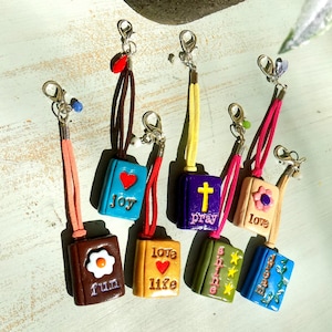 May include: A collection of seven miniature book charms with lobster clasps and suede cords. Each charm features a unique color and design: brown with 'fun', blue with 'joy', gold with 'love life', purple with 'pray', green with 'shine', beige with 'love', and blue with 'dream'.
