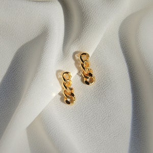May include: Two gold-colored chain link earrings. Each earring features a series of interconnected links, creating a textured, dangling design. The earrings are displayed against a soft, white fabric background, highlighting their metallic sheen.
