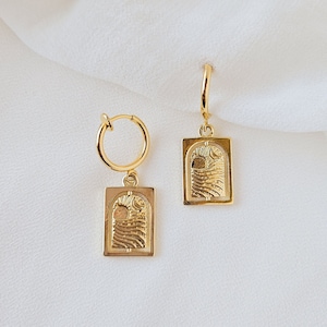Könnte beinhalten: Ein Paar goldene Creolen mit einem rechteckigen Anhänger, der ein strukturiertes Design aus Wellen und einer Sonne zeigt.