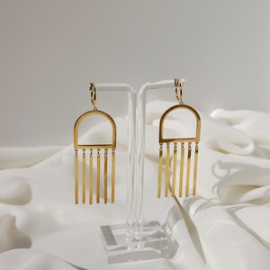May include: A pair of gold hoop earrings with a geometric arch design and dangling vertical bars. The earrings are displayed on a clear acrylic stand.