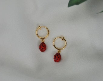 May include: A pair of gold hoop earrings with a red ladybug charm hanging from each hoop.