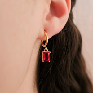 A small-sized dark ruby red cubic zirconia crystal emerald-cut rectangle charm with 18 karat gold plated border dangling from a small thin 18-karat-gold plated clip-on huggie hoop closure, which is hypoallergenic, nickel-free, and invisible acrylic.
