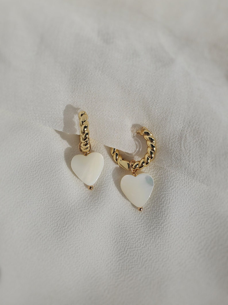 Medium-sized cream white heart-shaped natural freshwater pearl shells dangling from small 18-karat-gold plated twisted huggie hoops secured by hypoallergenic and nickel-free acrylic invisible clip-on closures.