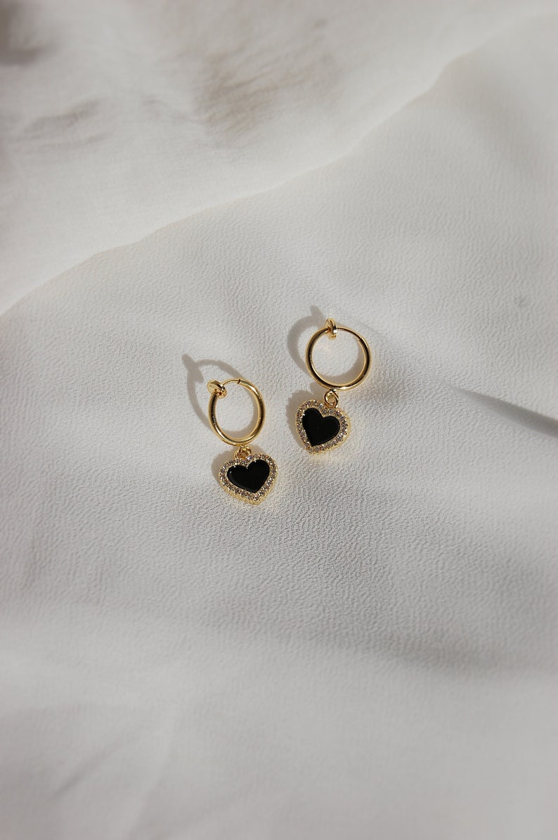 A single small 18-karat-gold plated and clear cubic zirconia crystal bordered black acrylic resin heart-shaped charm dangling from a small thin 18-karat-gold plated clip-on huggie hoop closure, which is hypoallergenic, nickel-free, and invisible.