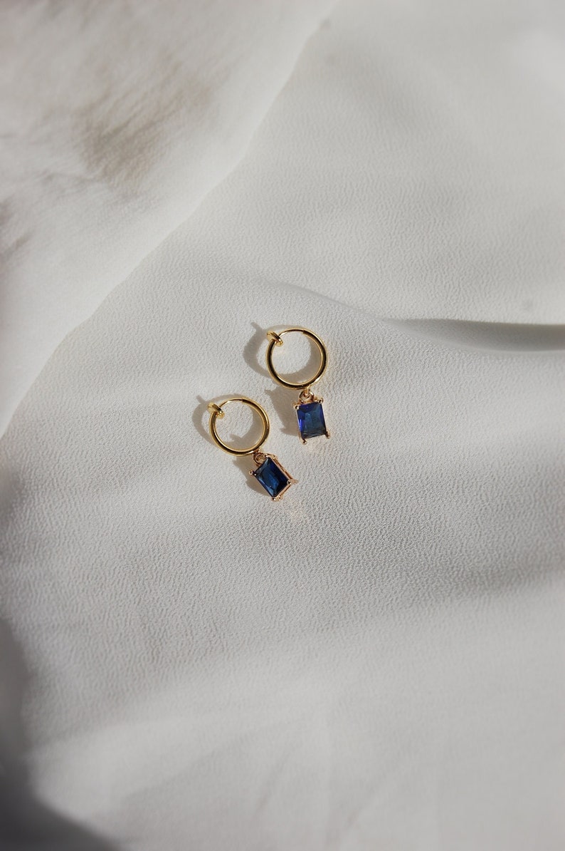 A small-sized sapphire blue cubic zirconia crystal emerald-cut rectangle charm with 18 karat gold plated border dangling from a small thin 18-karat-gold plated clip-on huggie hoop closure, which is hypoallergenic, nickel-free, and invisible acrylic.