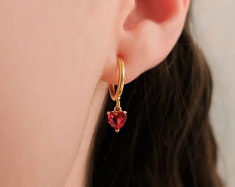 May include: A gold hoop earring with a small, heart-shaped red gemstone pendant. The earring is worn on an ear, with the focus on the jewelry's design and color. The earring's gold color contrasts with the red gemstone.