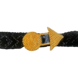 May include: A black braided leather belt with a gold and black buckle. The buckle is a round circle with a triangular point on one side. The buckle has a black stone in the centre.