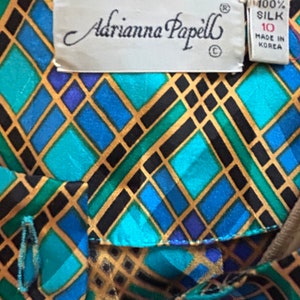 K&ouml;nnte beinhalten: Eine blaue, gr&uuml;ne und goldene geometrisch gemusterte Seidenbluse mit einem Etikett mit der Aufschrift "Adrianna Papell" und "100% Seide, hergestellt in Korea".