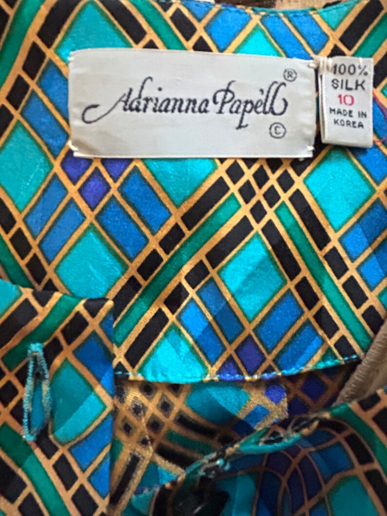 K&ouml;nnte beinhalten: Eine blaue, gr&uuml;ne und goldene geometrisch gemusterte Seidenbluse mit einem Etikett mit der Aufschrift "Adrianna Papell" und "100% Seide, hergestellt in Korea".