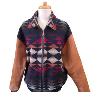 May include: A zip-up jacket featuring a dark navy blue body with a geometric diamond pattern in shades of red, orange, and tan. The jacket includes solid tan leather sleeves, a fold-down collar with the same geometric pattern, and ribbed cuffs.