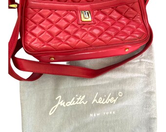May include: A red quilted leather crossbody bag with a gold-tone clasp. The bag has a long, adjustable strap and a matching red lining. A gray dust bag with the text "Judith Leiber New York" is included.