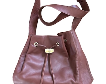 May include: A brown leather bucket bag with a long, adjustable shoulder strap. The bag features a drawstring closure and a gold-toned metal accent on the front. The bag's dimensions are approximately 30 cm wide, 25 cm high, and 20 cm deep.