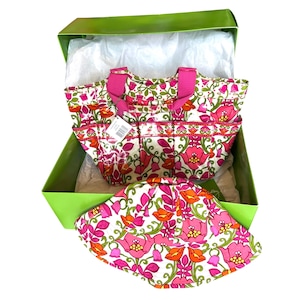 Puede incluir: Un conjunto de bolso tote y sombrero para el sol con estampado floral en una caja verde. El bolso tiene asas y bolsillos rosas, y el sombrero tiene un patrón floral similar con acentos rosas, naranjas y verdes. El conjunto se presenta sobre papel de seda blanco.