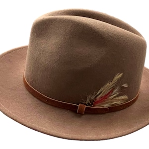 Puede incluir: Un sombrero fedora de fieltro marrón con una banda de cuero marrón y un pequeño adorno de plumas.