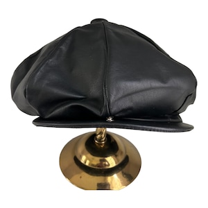 May include: A black leather newsboy cap with a rounded crown and a brim. The cap is displayed on a gold stand.