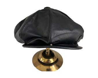 Könnte beinhalten: Eine schwarze Leder-Newsboy-Mütze mit abgerundeter Krone und einem Schirm. Die Mütze wird auf einem goldenen Ständer präsentiert.